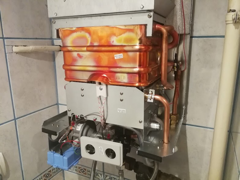 Otwarty piec gazowy z widocznym wymiennikiem ciepła z miedzianymi rurami, zamontowany na ścianie wyłożonej kafelkami.