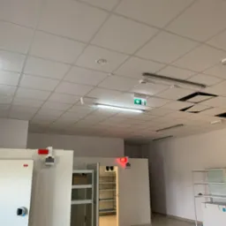 Sufit kasetonowy z oświetleniem LED, czujnikiem dymu i znakiem ewakuacyjnym. Widoczne niedokończone prace i drabina. Oświetlenie magazynowe.