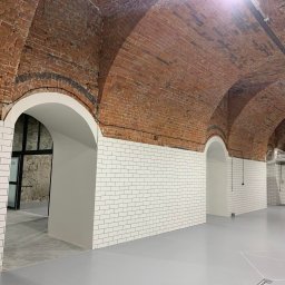 Instalacje elektryczne Krzysztof Niemiec - Wnętrze z ceglanym sklepieniem łukowym, białymi płytkami ściennymi i szarą podłogą. Widoczne przewody elektryczne pod sufitem i elementy wentylacji.