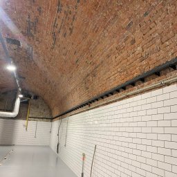 Instalacje elektryczne Krzysztof Niemiec - Wnętrze po remoncie z ceglanym sklepieniem i białymi kafelkami. Widoczna instalacja kablowa, rury i akcesoria, co sugeruje prace elektryczne w trakcie lub po instalacji.