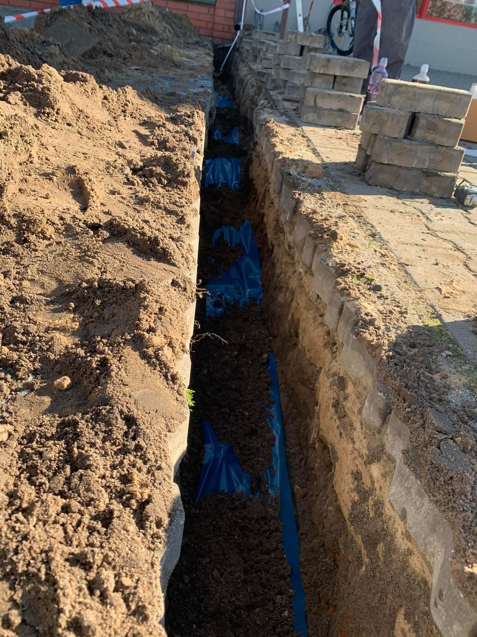 Wkopany rów z niebieską folią ochronną, przygotowany pod instalację, z kostką brukową w tle. Prace ziemne przy budynku.
