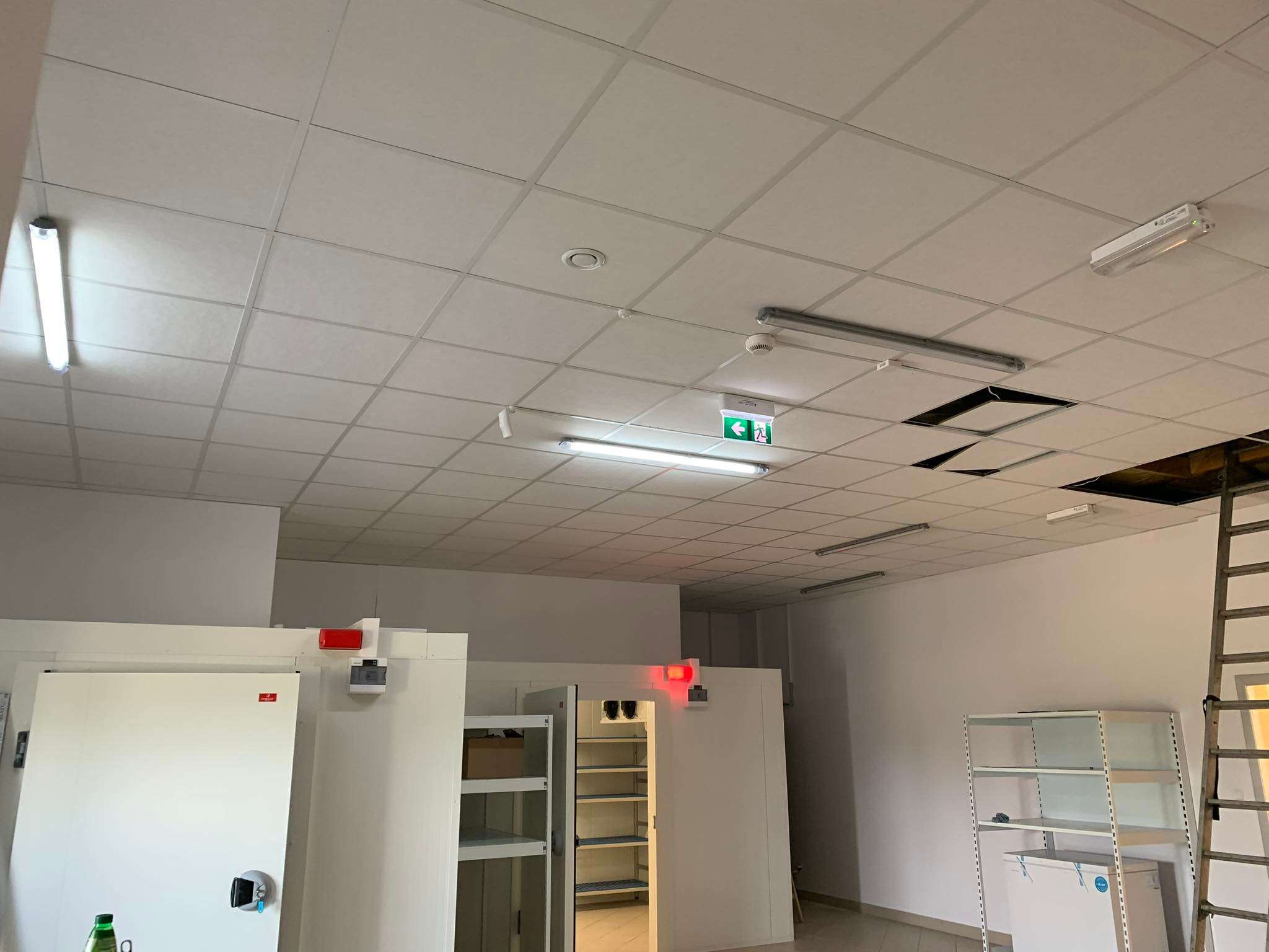 Sufit kasetonowy z oświetleniem LED, czujnikiem dymu i znakiem ewakuacyjnym. Widoczne niedokończone prace i drabina. Oświetlenie magazynowe.