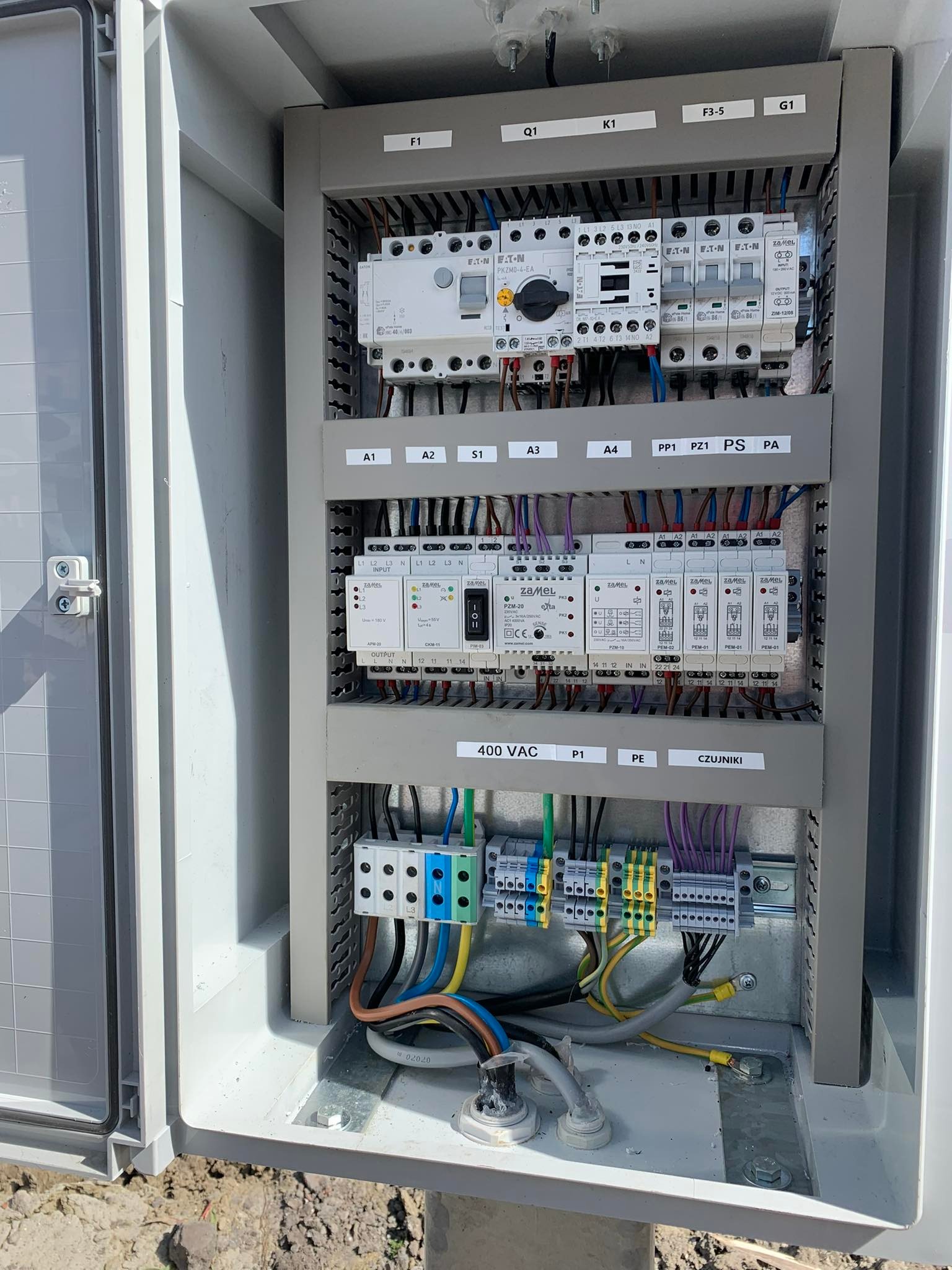 Otwarta szara szafa sterownicza z widocznymi bezpiecznikami, przewodami i modułami Zamel. Elementy oznaczone etykietami, widoczne złącza kablowe i przewody.