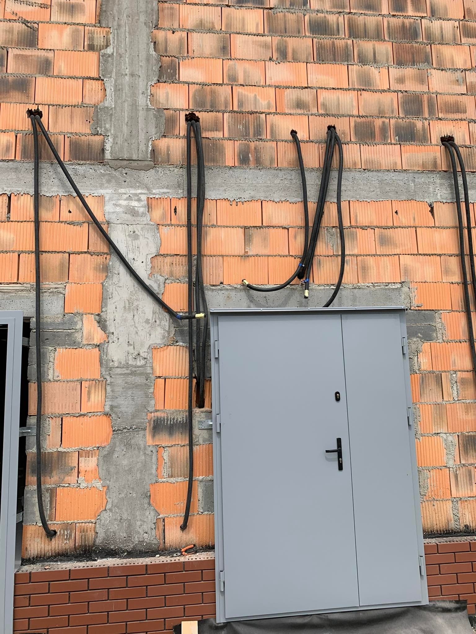 Czarne rury w karbowanej osłonie przymocowane do surowej ściany z cegły dziurawki, nad szarymi drzwiami wejściowymi. Etap instalacji.
