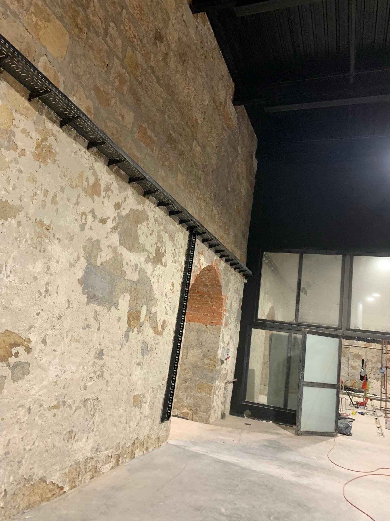 Metalowa trasa kablowa zamontowana na starej, kamiennej ścianie w surowym wnętrzu z oknami. Widoczne ślady remontu i elementy konstrukcyjne.