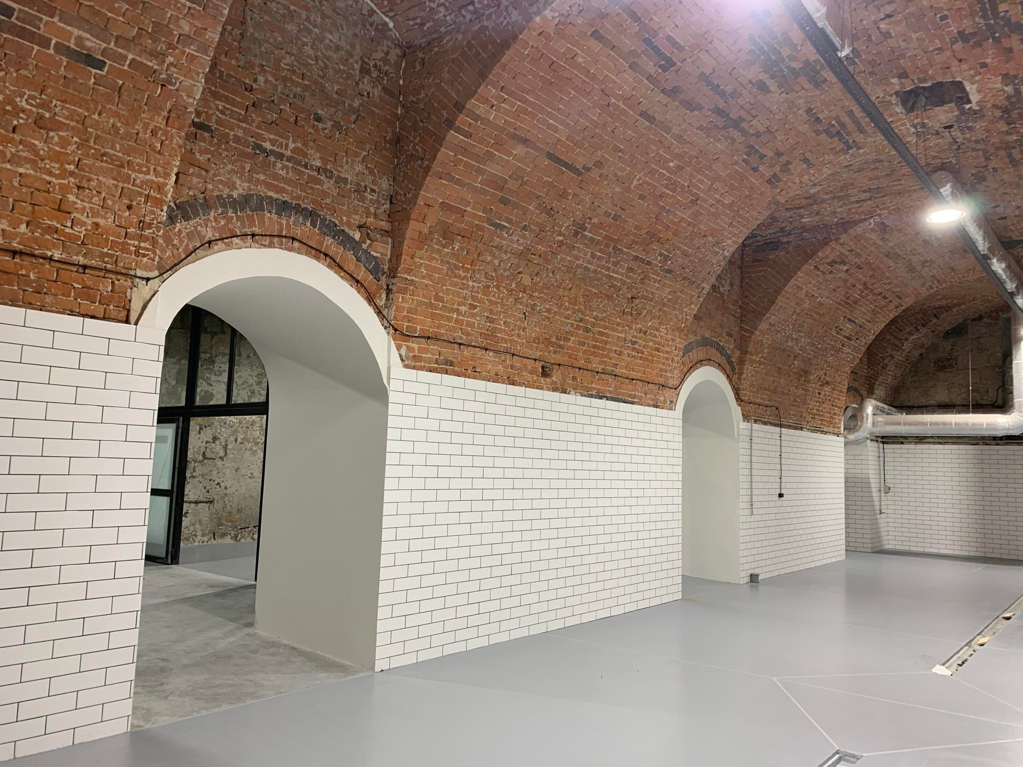 Wnętrze z ceglanym sklepieniem łukowym, białymi płytkami ściennymi i szarą podłogą. Widoczne przewody elektryczne pod sufitem i elementy wentylacji.