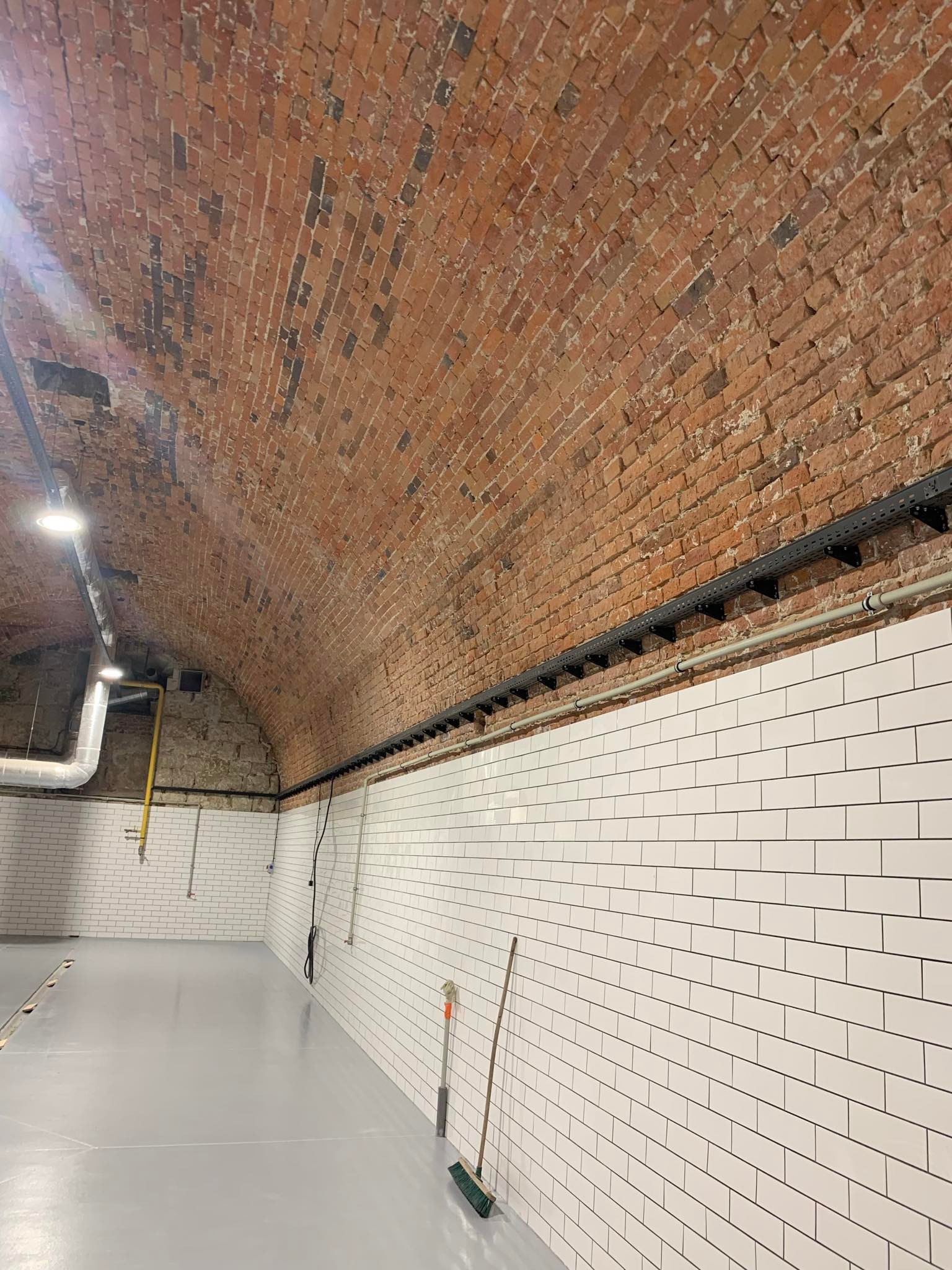 Wnętrze po remoncie z ceglanym sklepieniem i białymi kafelkami. Widoczna instalacja kablowa, rury i akcesoria, co sugeruje prace elektryczne w trakcie lub po instalacji.