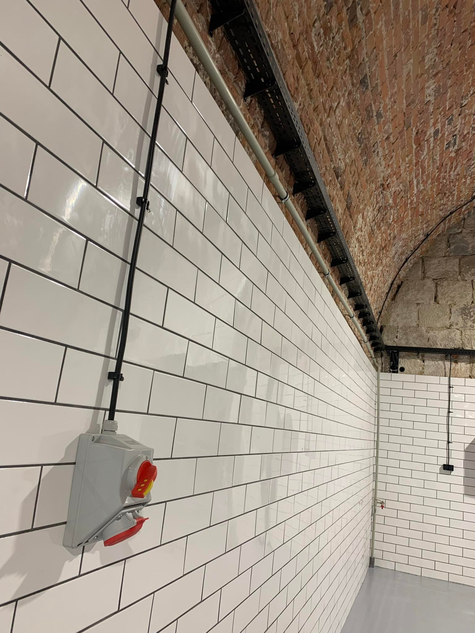 Szara skrzynka elektryczna z czerwonymi elementami na białej, kafelkowanej ścianie. Widoczne przewody i koryto kablowe pod ceglanym sklepieniem.