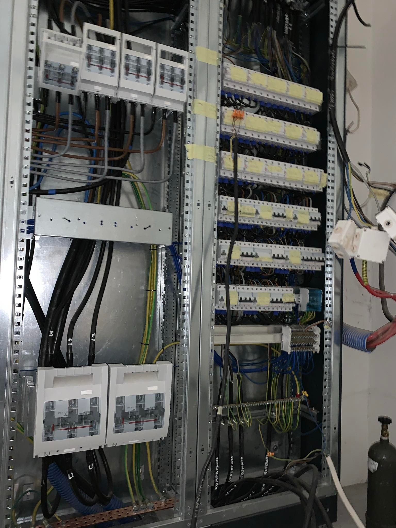 Rozdzielnica elektryczna z licznymi przewodami, bezpiecznikami i listwami zaciskowymi, w metalowej obudowie. Widoczne kable oznaczone etykietami.