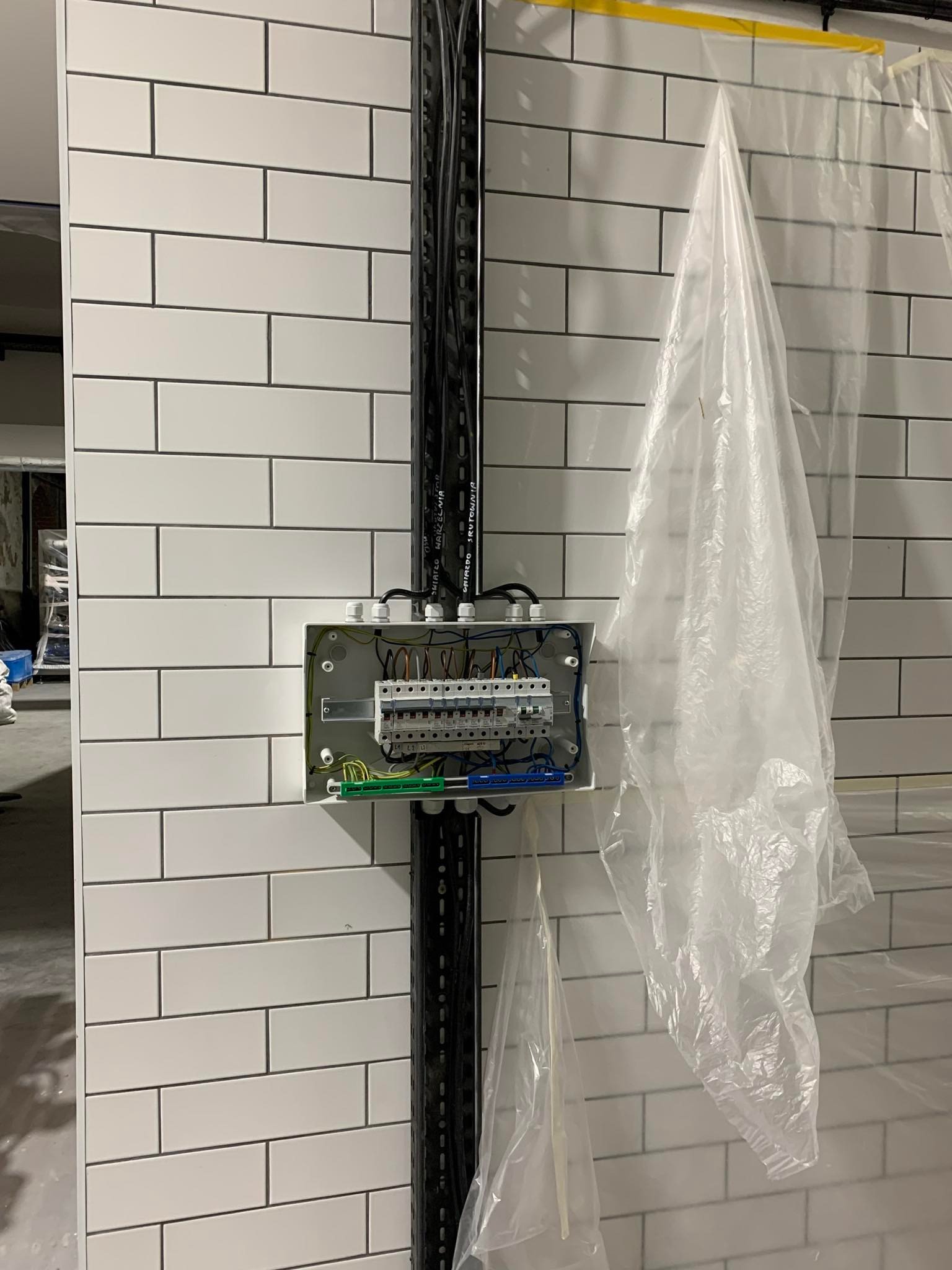 Szafka elektryczna z przewodami zamontowana na ścianie z białych płytek, zabezpieczona folią malarską. Widoczna metalowa szyna z kablami.