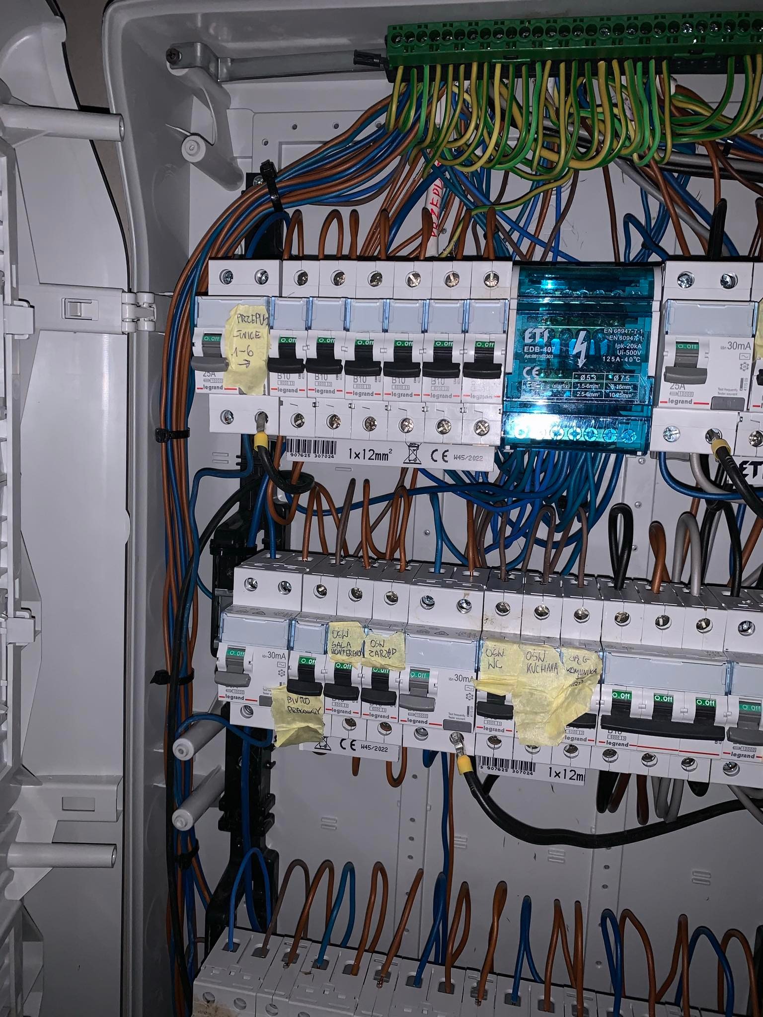 Rozdzielnia elektryczna z bezpiecznikami Legrand, okablowaniem i naklejonymi etykietami. Widoczne przewody w różnych kolorach i aparatura modułowa.