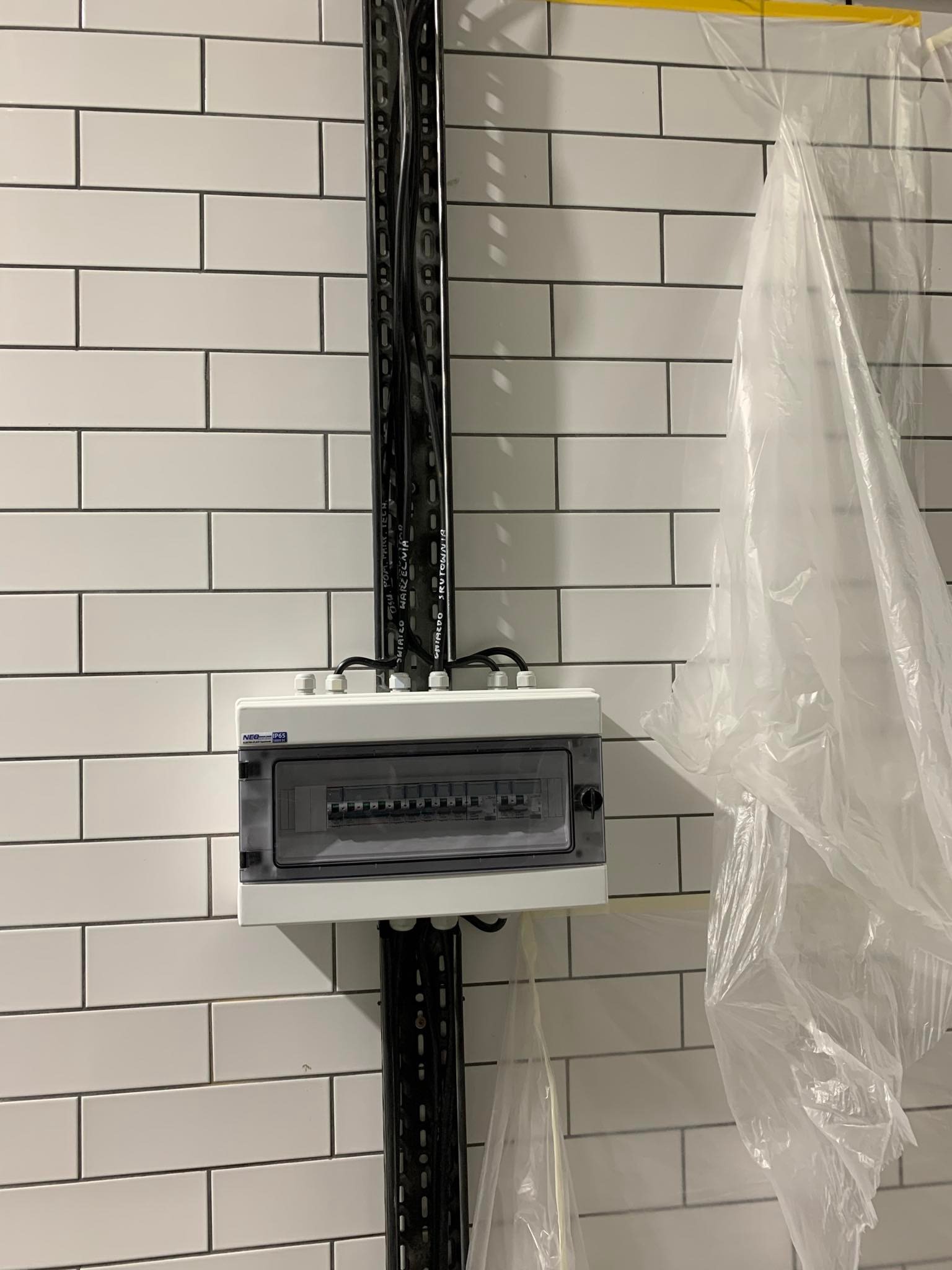 Zamontowana skrzynka rozdzielcza z przewodami na ścianie wyłożonej białymi płytkami, zabezpieczona folią malarską. Widoczne czarne kable w metalowej rynnie.