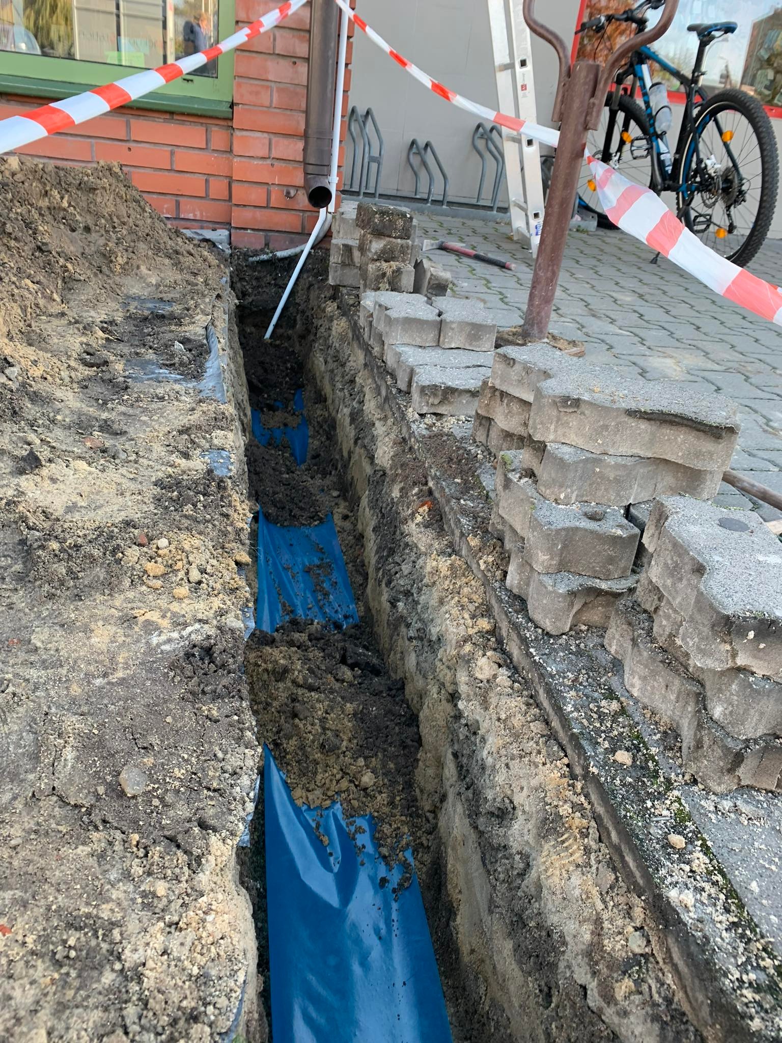 Wykop w ziemi zabezpieczony folią, z widocznymi kablami. Prace ziemne w pobliżu budynku z cegły, oznaczone taśmą ostrzegawczą. Widoczne elementy infrastruktury.