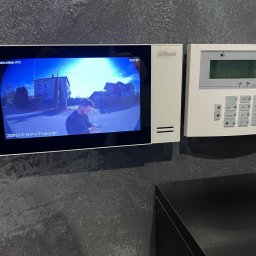 SWS Instal - Zestaw alarmowy Satel obok wideodomofonu Dahua z podglądem na ulicę, zamontowane na teksturowanej, ciemnej ścianie. Widoczny fragment czarnego mebla.