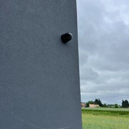 SWS Instal - Czarna kamera Dahua zamontowana na szarej, teksturowanej ścianie budynku z widokiem na zielone pole i pochmurne niebo w tle. Obraz z bliska.
