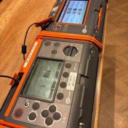 SWS Instal - Elektryk - Miernik instalacji elektrycznych Sonel MPI-520 na drewnianym blacie. Widoczne ekrany, przyciski, oznaczenia i przewody. Urządzenie w szarej obudowie z pomarańczowymi elementami.