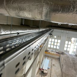 SWS Instal - Metalowa konstrukcja pod sufitem hali przemysłowej, prawdopodobnie trasa kablowa. Widoczne rury wentylacyjne i drzwi hali w tle. Ujęcie z dołu, perspektywa żabia.