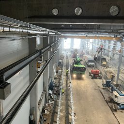 SWS Instal - Hala przemysłowa w trakcie instalacji. Widoczne koryta kablowe pod sufitem, maszyny budowlane i pracownicy. Ujęcie z góry, podkreślające rozległość przestrzeni.
