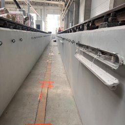 SWS Instal - Hala przemysłowa w trakcie instalacji oświetlenia: widoczne koryto kablowe, uchwyty i częściowo zamontowana oprawa. W tle materiały izolacyjne.