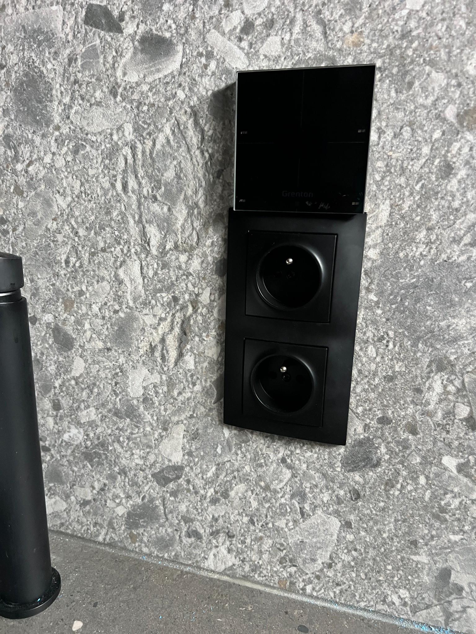 Dwa czarne gniazdka elektryczne zamontowane na szarej ścianie z fakturą kamienia, nad nimi panel sterowania inteligentnego domu marki Grenton.