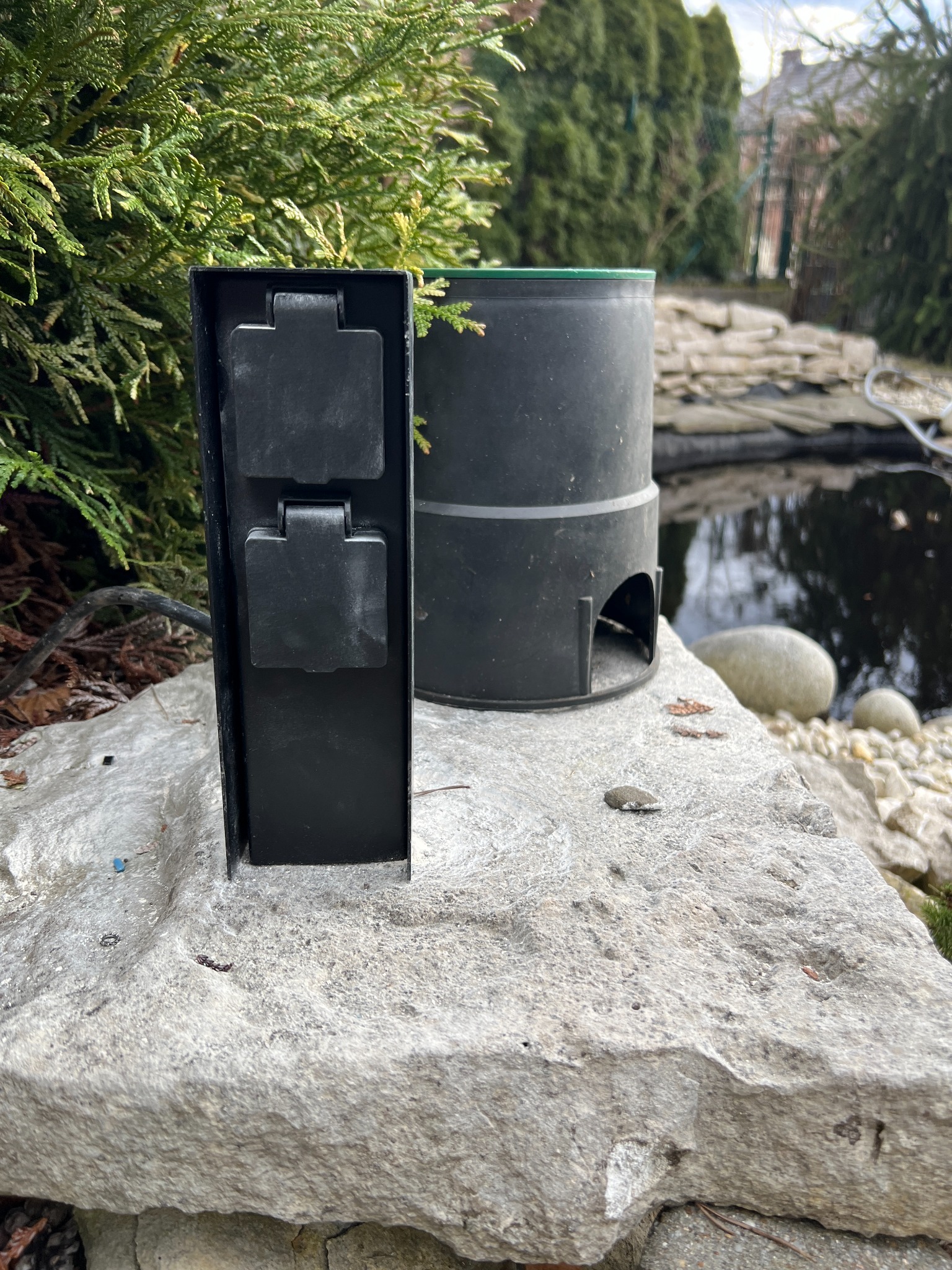 Zewnętrzna skrzynka elektryczna z dwoma gniazdami, umieszczona na kamieniu obok oczka wodnego i filtra, otoczona zielenią.