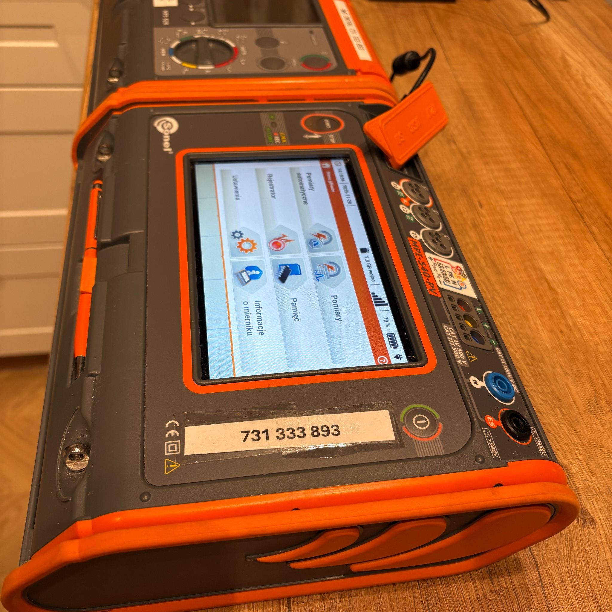 Miernik Sonel MPI-540-PV na drewnianej powierzchni. Urządzenie do pomiarów elektrycznych z widocznym ekranem i przyciskami. Numer kontaktowy na obudowie.