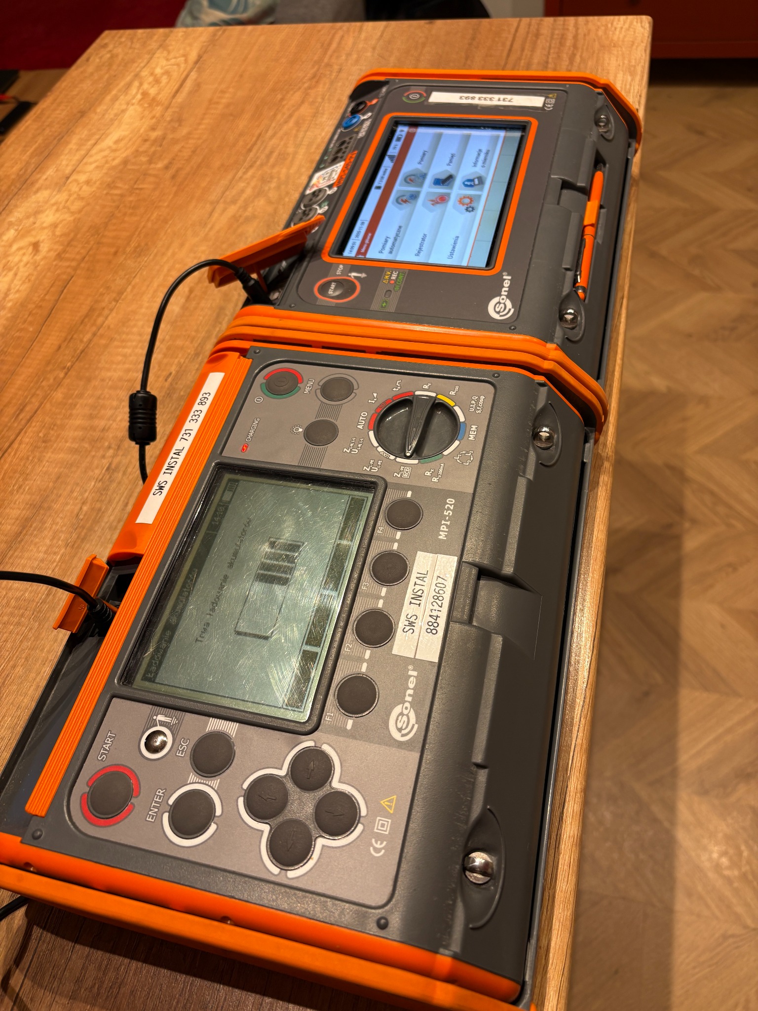 Miernik instalacji elektrycznych Sonel MPI-520 na drewnianym blacie. Widoczne ekrany, przyciski, oznaczenia i przewody. Urządzenie w szarej obudowie z pomarańczowymi elementami.