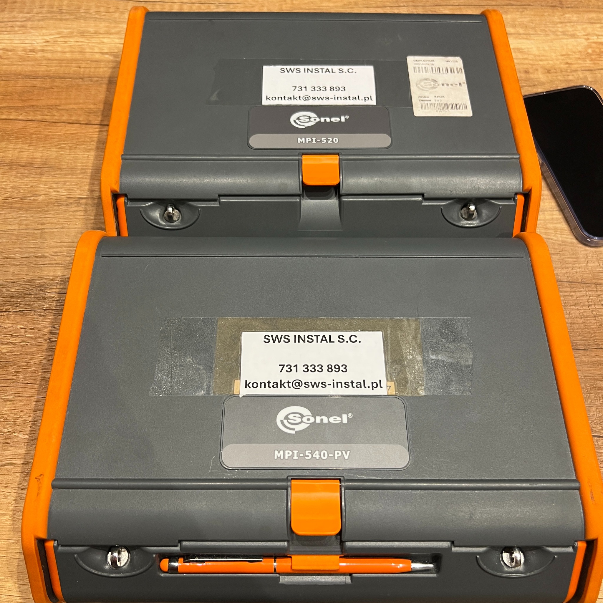 Dwa mierniki Sonel MPI-520 i MPI-540-PV z danymi firmy SWS Instal S.C., leżące jeden na drugim na drewnianym blacie, obok widoczny fragment smartfona.