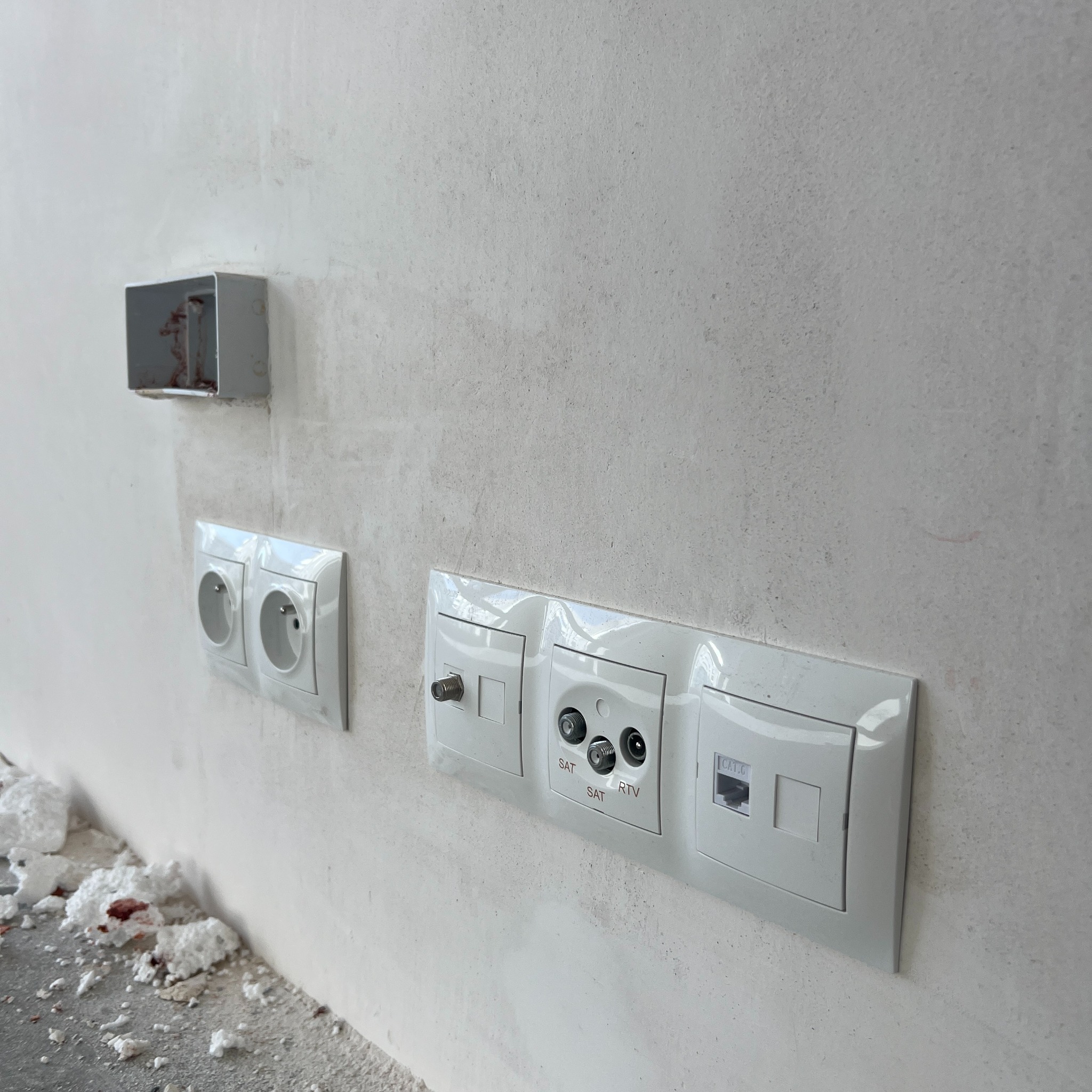 Białe gniazda elektryczne i antenowe (SAT, RTV, CAT.6) zamontowane na surowej, jasnej ścianie. Widoczna puszka instalacyjna i resztki styropianu na podłodze.