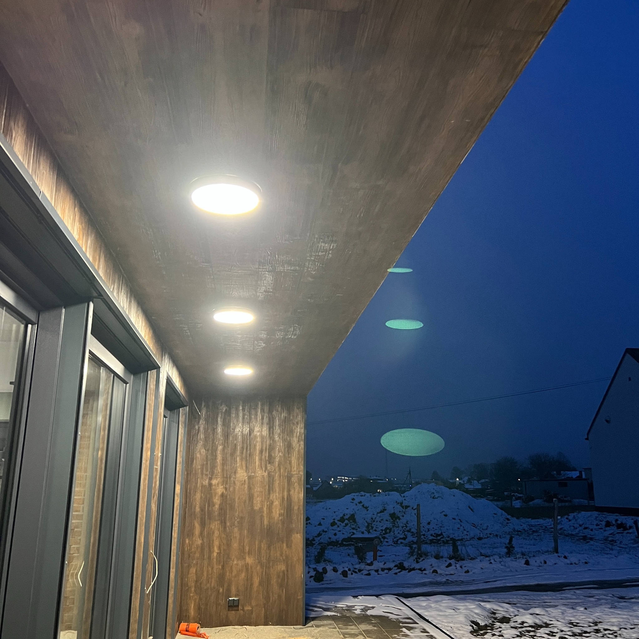Elewacja budynku z zamontowanymi, okrągłymi lampami LED, oświetlającymi teren. Widoczne okna i fragment zimowego krajobrazu w tle.