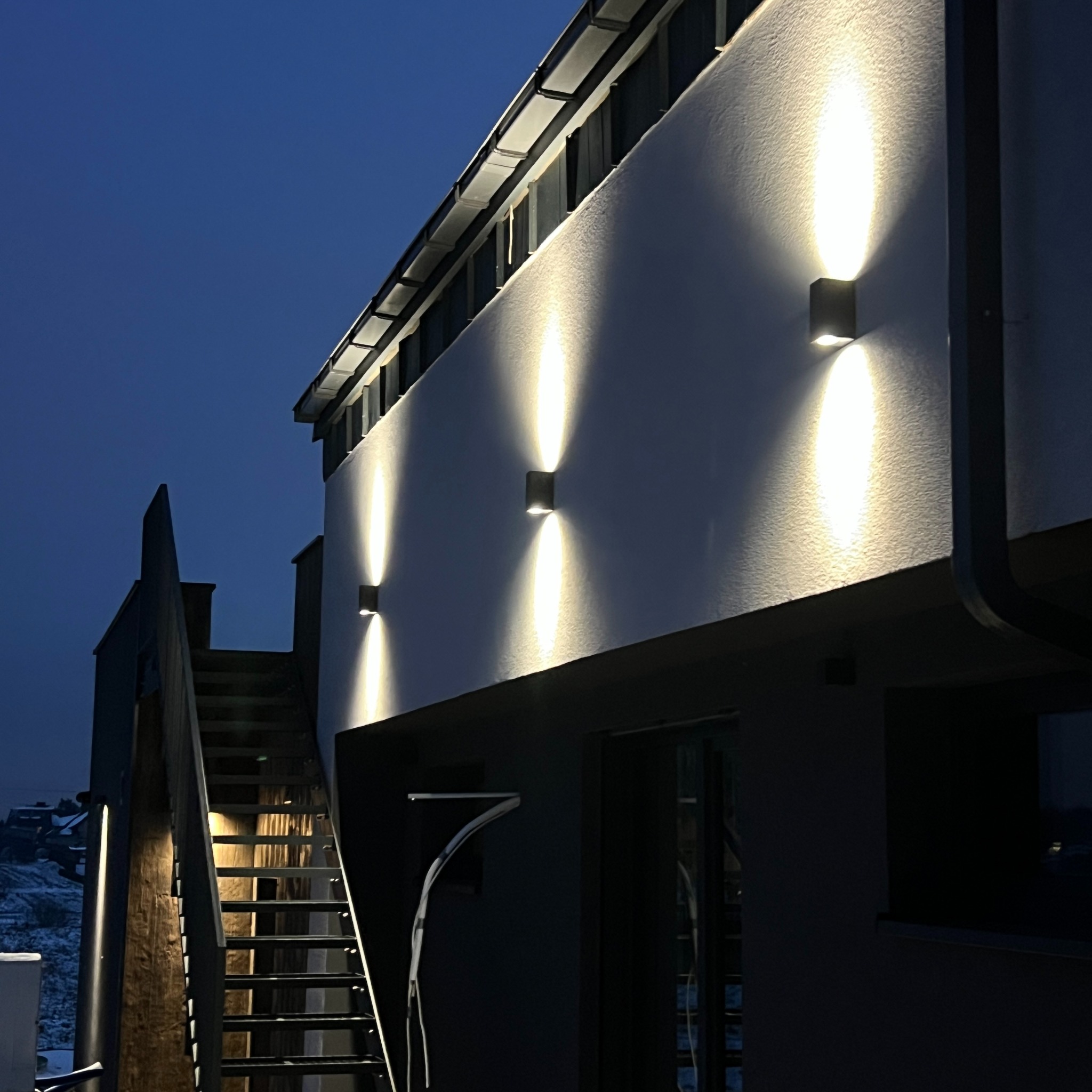 Elewacja budynku z zamontowanymi, świecącymi lampami zewnętrznymi, oświetlającymi ścianę. Widoczne schody zewnętrzne i fragment krajobrazu w tle.