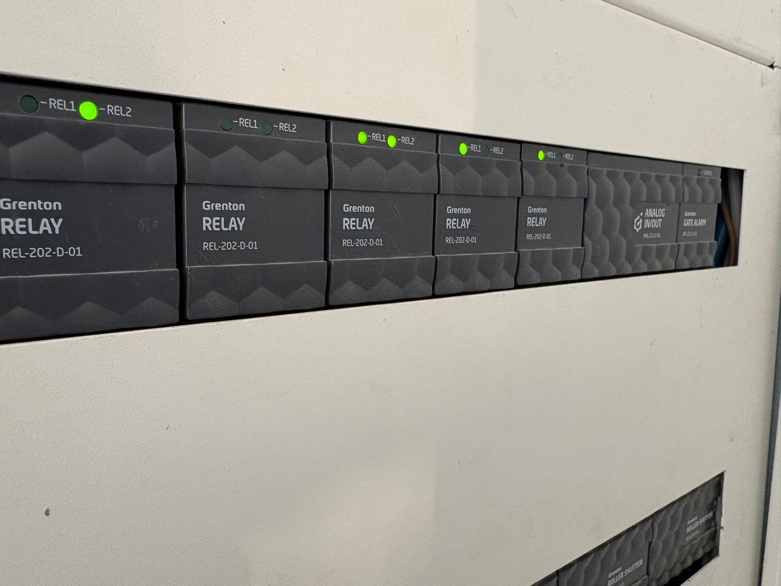 Panel sterowania inteligentnego domu Grenton z modułami przekaźnikowymi i analogowymi, diody LED sygnalizują stan pracy. Widoczne oznaczenia REL-202-D-01 i inne.