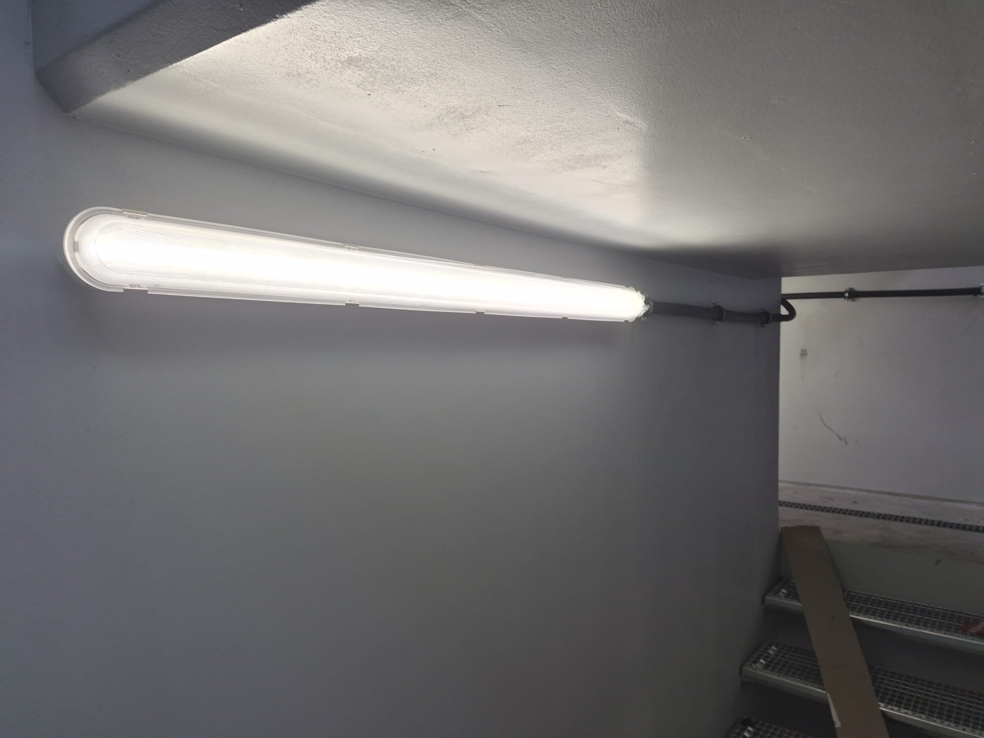 Zamontowana podłużna lampa przemysłowa LED na szarej ścianie w hali, widoczne rury instalacyjne i schody w tle. Oświetlenie sufitowe w obiekcie przemysłowym.