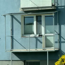 Zadaszony balkon z metalową konstrukcją, widok z dołu na tle niebieskiej elewacji budynku i zieleni.