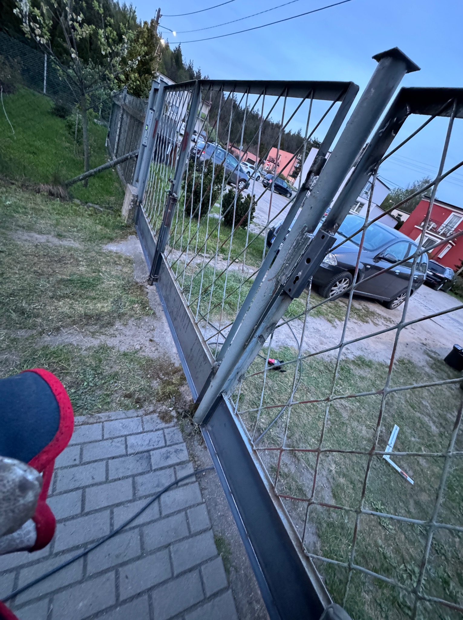 Metalowa brama w trakcie montażu, widoczne świeże spawy, tło z domami i samochodami, perspektywa z dołu.