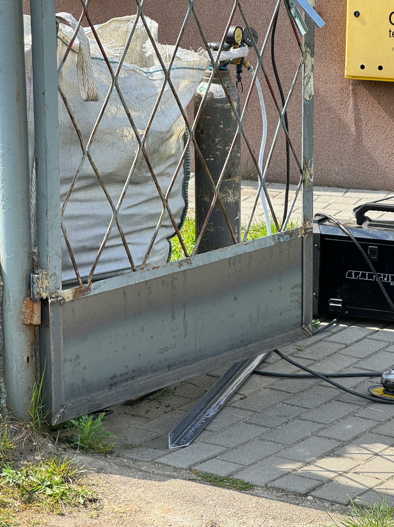 Fragment metalowej bramy w trakcie naprawy, widoczny spawany profil, butla z gazem, spawarka i szlifierka kątowa na brukowanej nawierzchni.