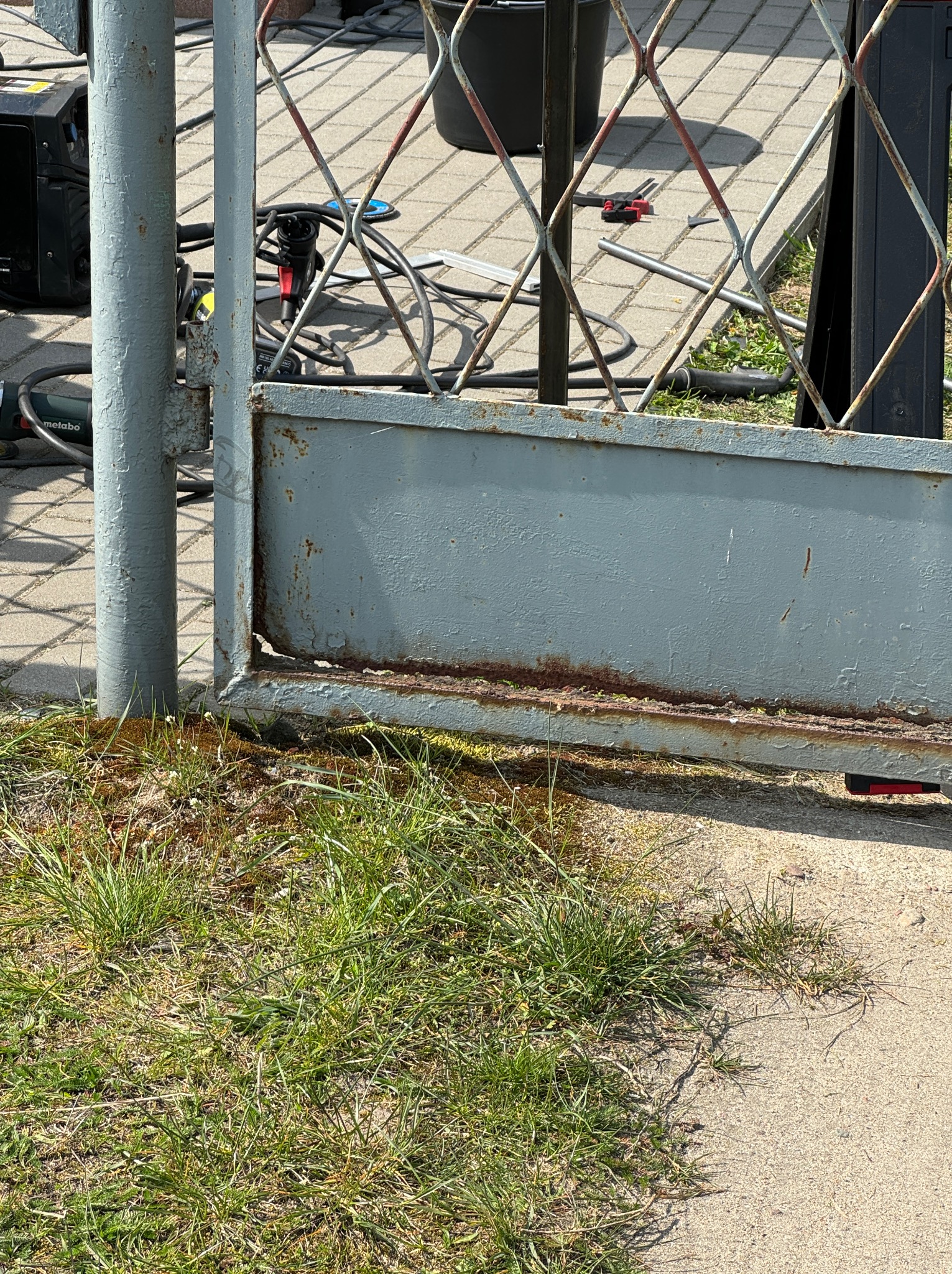 Fragment metalowej bramy z widocznymi śladami korozji u podstawy, trawa i narzędzia w tle.