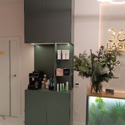 Studio mebli FABIANI - Nowoczesna zabudowa meblowa w salonie kosmetycznym: ciemnozielone szafki z eksposem na kawę, podświetlane półki z kosmetykami, marmurowa podłoga i dekoracje.