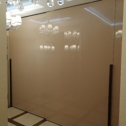 Studio mebli FABIANI - Beżowa szafa wnękowa z lustrzanymi drzwiami przesuwnymi i długimi, pionowymi uchwytami, odbijająca kryształowy żyrandol w eleganckim wnętrzu.