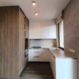 Studio mebli FABIANI - Nowoczesna kuchnia w odcieniach bieli i drewna. Zabudowa kuchenna z piekarnikiem, płyta indukcyjna i szary blat. Okno z roletą rzymską. Minimalistyczny design.