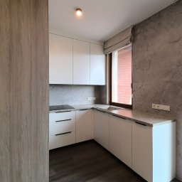 Studio mebli FABIANI - Nowoczesna, narożna kuchnia na wymiar z białymi frontami, czarnymi uchwytami i blatem imitującym marmur. Widok na okno z roletą rzymską i szarą ścianę.