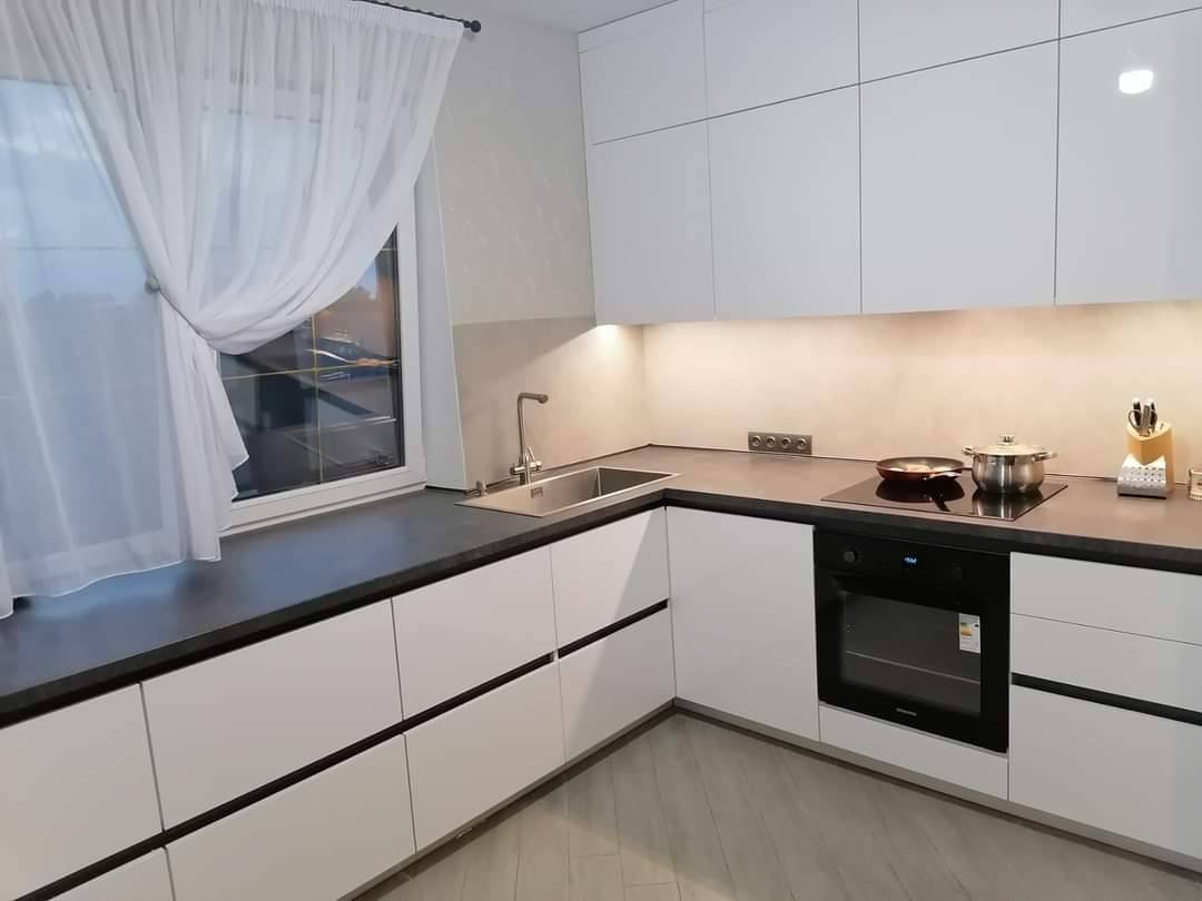 Nowoczesna kuchnia w kształcie litery L z białymi frontami szafek i ciemnym blatem, widok na okno z delikatną firanką, płyta indukcyjna z patelnią i garnkiem.