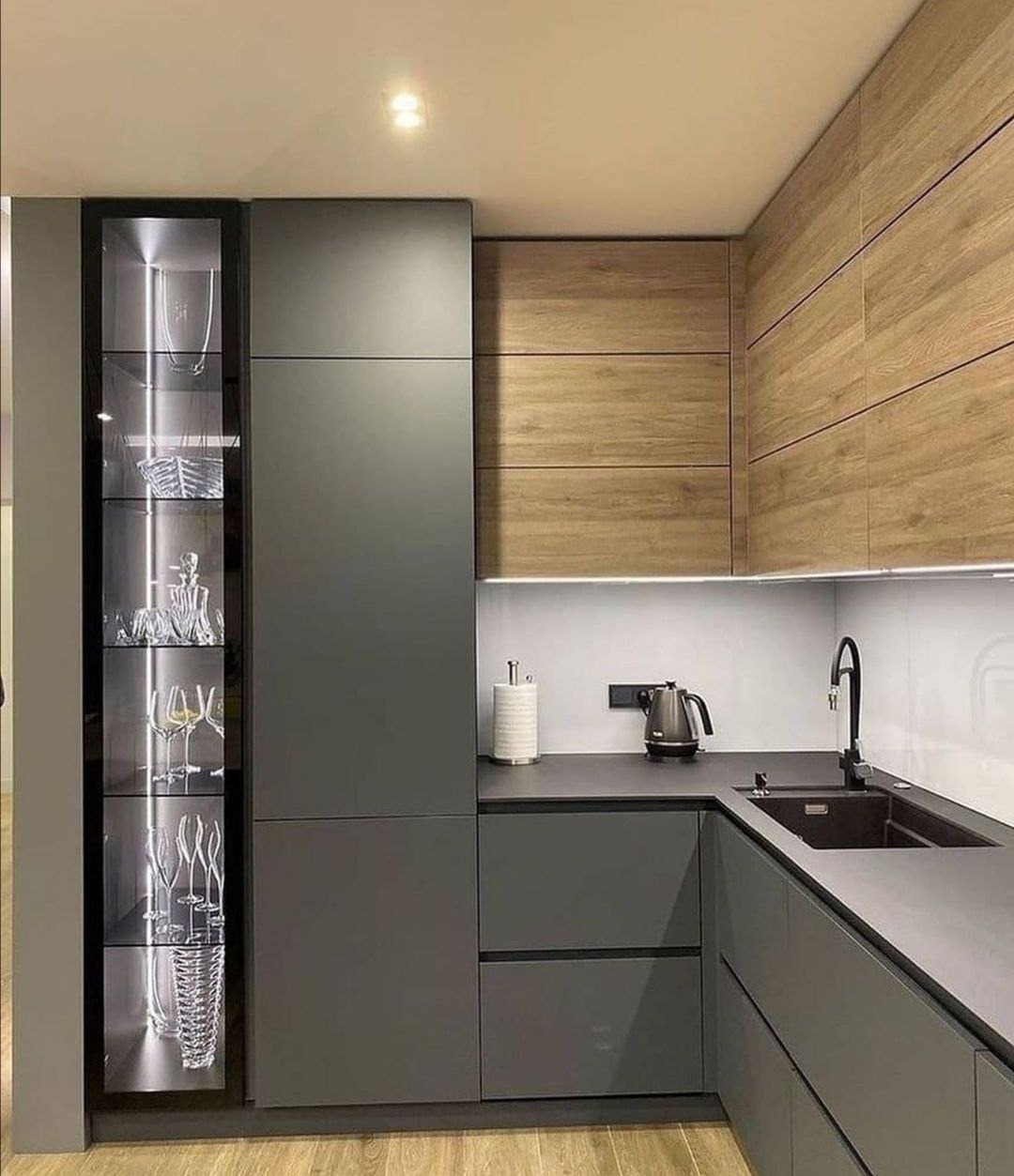 Nowoczesna kuchnia w kształcie litery L z szarymi szafkami i drewnianymi frontami górnymi, oświetlona listwą LED pod szafkami, z przeszkloną witryną eksponującą kryształowe naczynia.