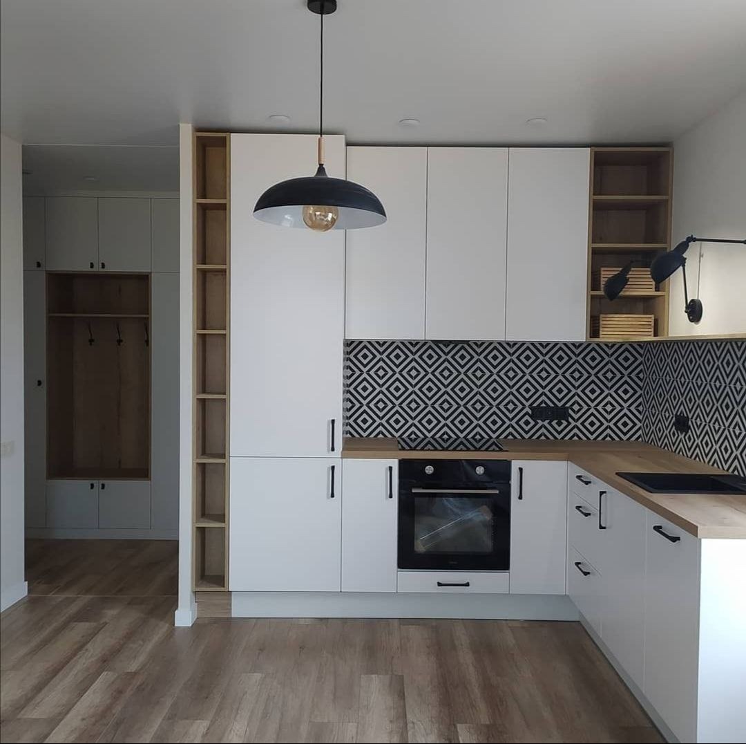 Nowoczesna kuchnia w kształcie litery L z białymi frontami, drewnianym blatem i wzorzystą czarno-białą mozaiką nad blatem, oświetlona wiszącą lampą i kinkietem.