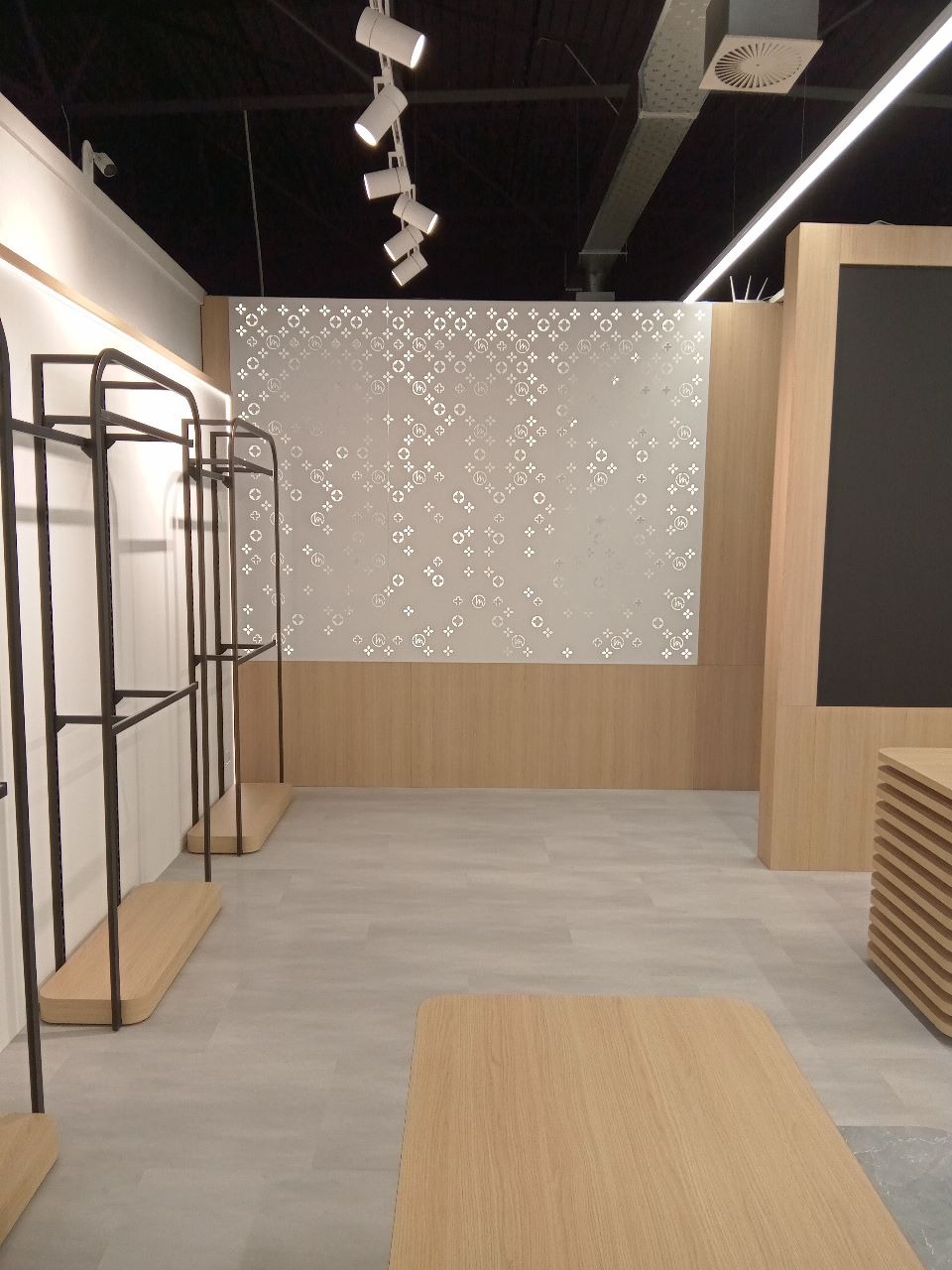 Nowoczesne wnętrze sklepu odzieżowego z drewnianymi elementami, metalowymi wieszakami i oświetleniem punktowym. Minimalistyczny design z wzorzystą ścianą w tle.