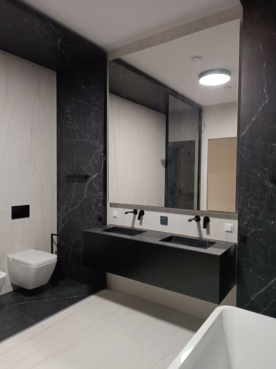 Nowoczesna łazienka z czarną szafką pod umywalką i marmurowymi płytkami. Minimalistyczny design, podwieszana toaleta i duże lustro.