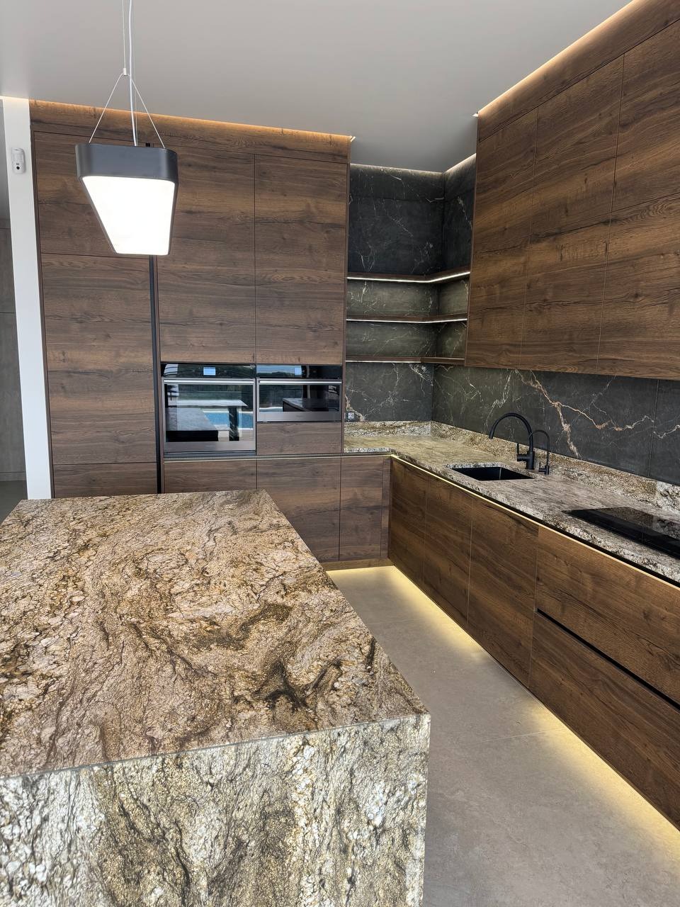 Nowoczesna kuchnia z drewnianymi frontami, blatem z kamienia naturalnego i podświetleniem LED. Widoczna wyspa kuchenna i zabudowa z piekarnikiem.