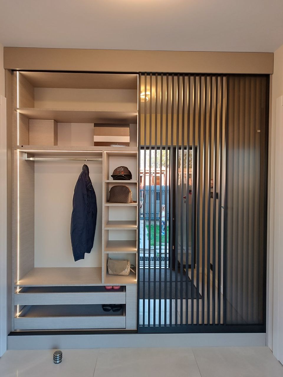 Jasna szafa wnękowa z przesuwnymi drzwiami z pionowych listew, oświetleniem LED i wyposażeniem wewnętrznym: półki, szuflady, drążek na ubrania. Widok na wnętrze domu.