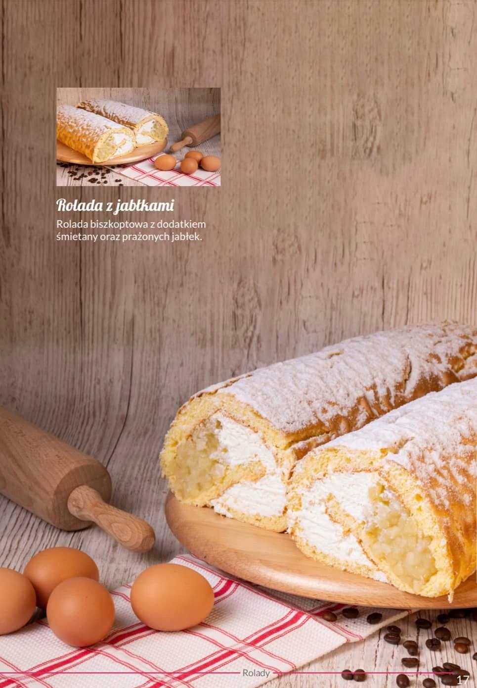 Biszkoptowa rolada z jabłkami i kremem śmietankowym, posypana cukrem pudrem, leżąca na drewnianej desce obok jajek i drewnianego wałka na tle drewnianych desek.
