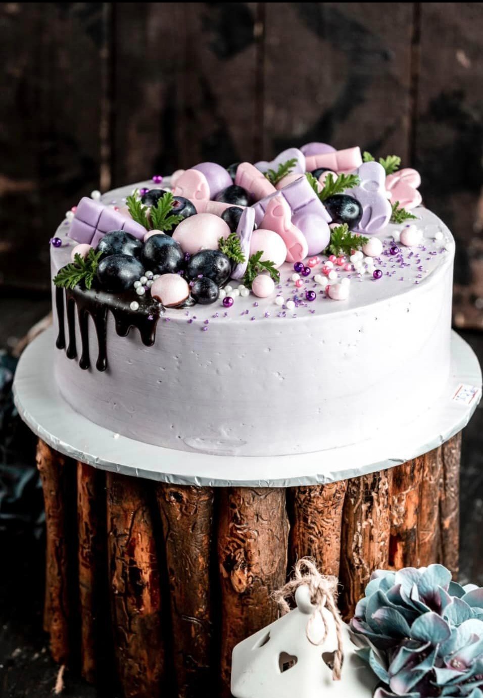 Tort z jasnofioletowym kremem, udekorowany borówkami, czekoladową polewą i pastelowymi ozdobami z masy cukrowej, umieszczony na rustykalnym drewnianym stojaku.